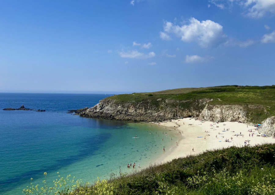 Paysage du littoral breton sous le soleil. On y voit une plage de sable blanc bordée d'une mer bleu turquoise